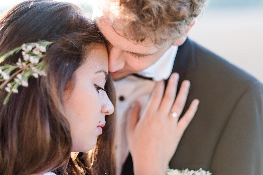 Ein Paar umarmt sich; der Mann küsst die Frau auf die Stirn. Die Frau trägt einen Blumenkranz, beide haben die Augen geschlossen.