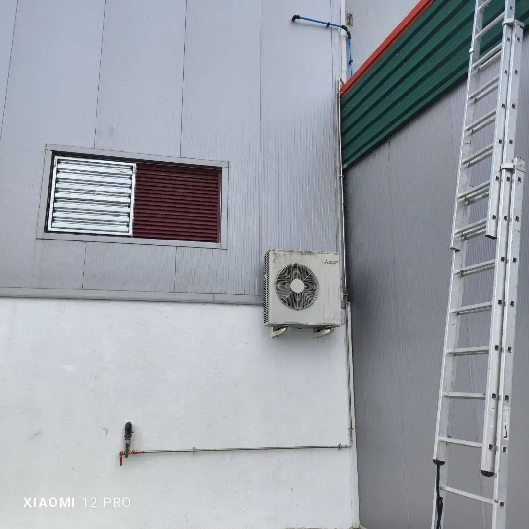Pared exterior con una ventana, una unidad de aire acondicionado y una escalera junto a un edificio de chapa ondulada.