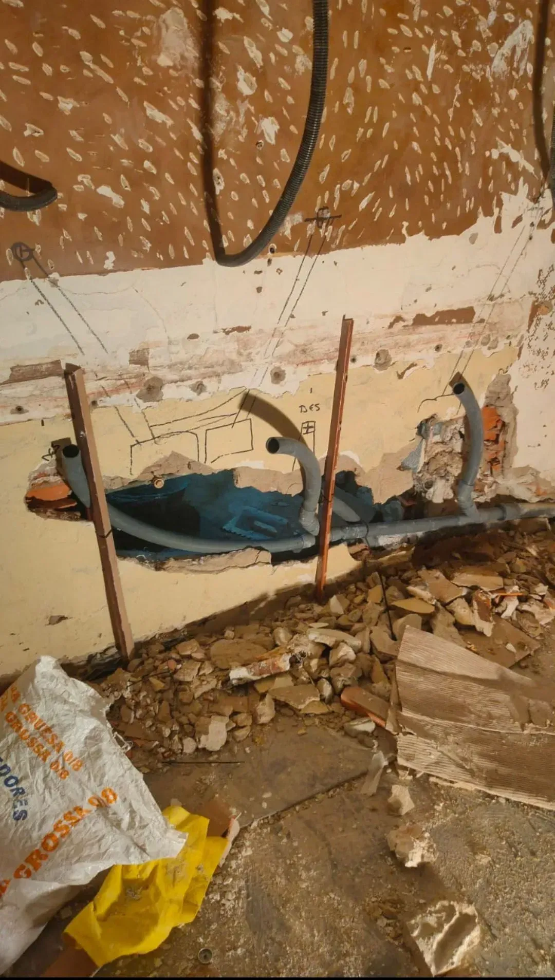Pared dañada con tuberías expuestas, yeso roto y escombros en el suelo dentro de una habitación.