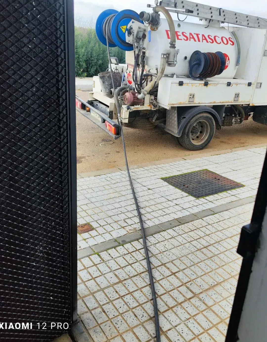 Un camión de alcantarillado blanco con mangueras azules estacionado en una calle embaldosada y mojada, detrás de una valla.