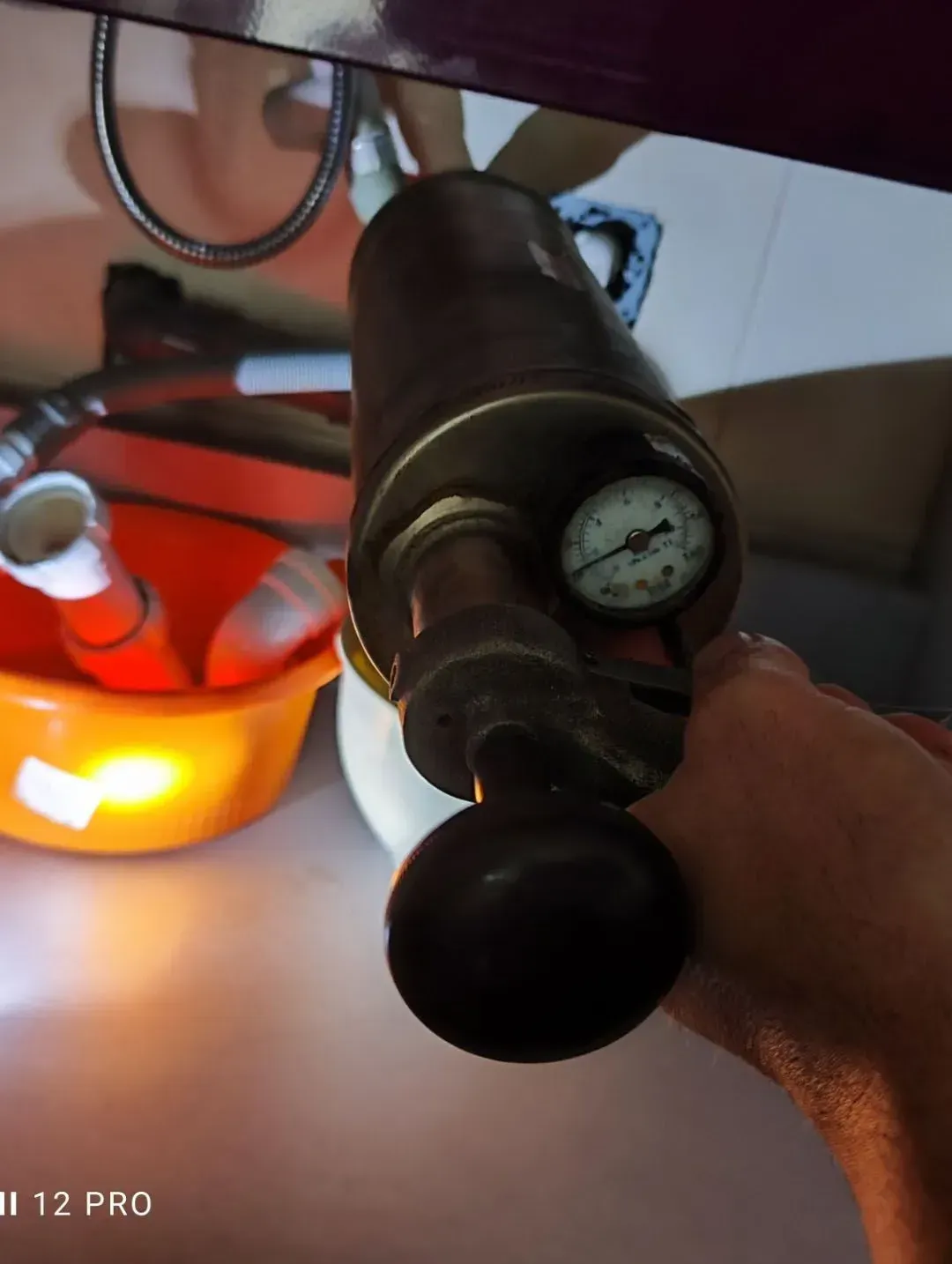 Primer plano de una mano que sostiene un dispositivo metálico con un indicador redondo y una perilla negra, iluminado por una lámpara cálida.