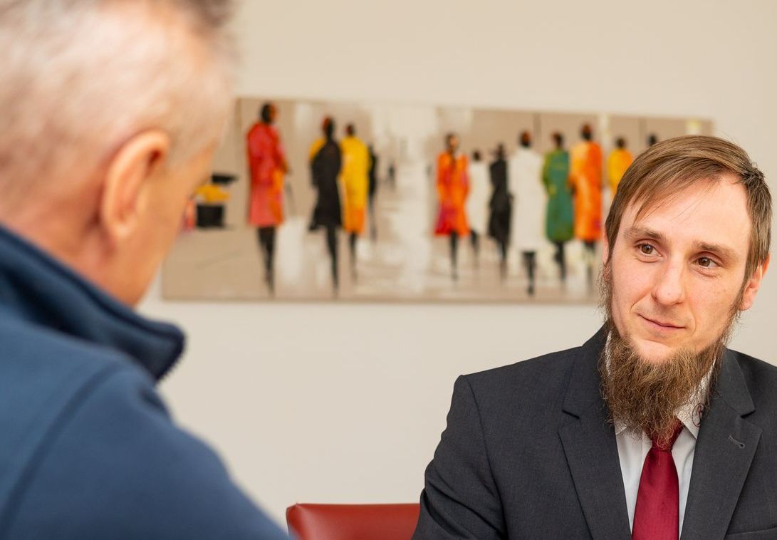 Ein Mann in Anzug und Krawatte sitzt an einem Tisch und unterhält sich mit einem anderen Mann