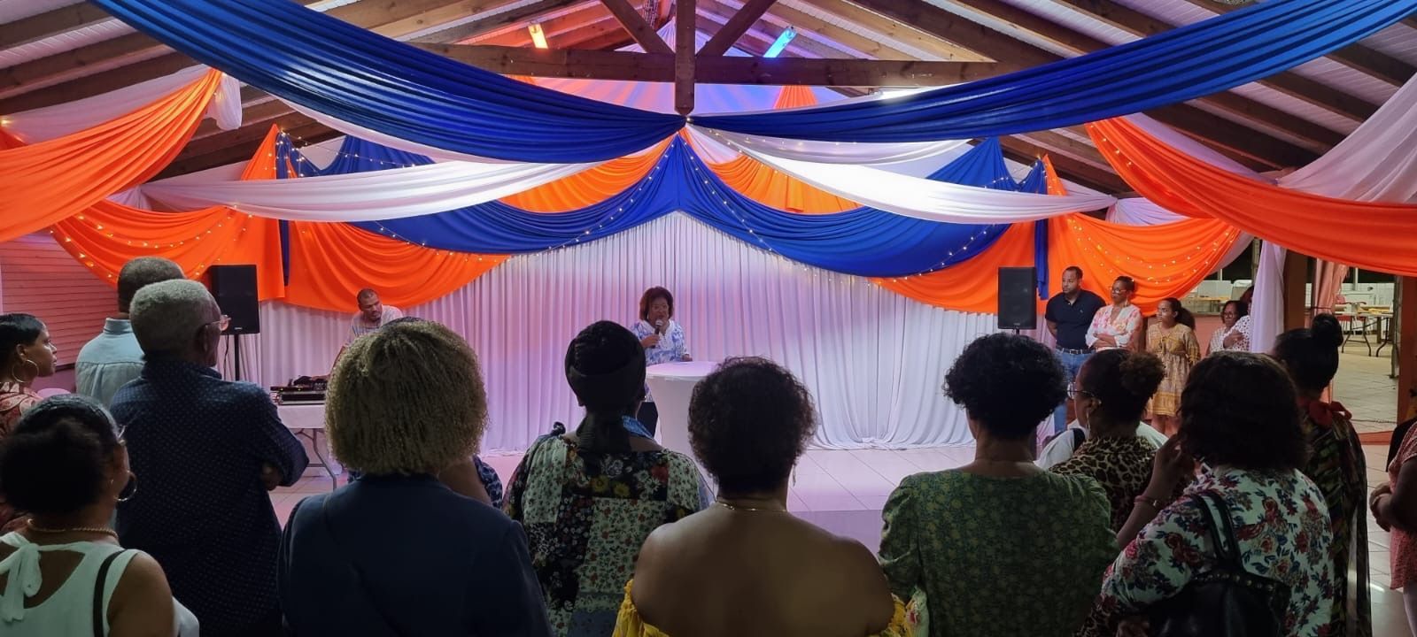 Un groupe de personnes se tient debout dans une salle décorée de tissus orange, bleus et blancs, face à un orateur à la tribune.