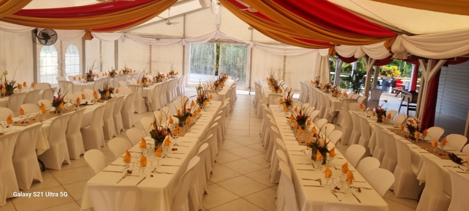 Une tente aménagée pour les banquets, avec de longues tables recouvertes de nappes blanches et décorée de tentures de plafond dorées et rouges.