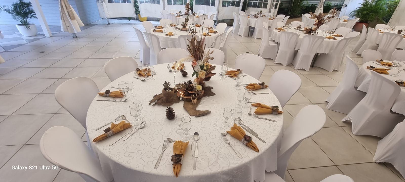 Des tables rondes dans une salle de réception lumineuse, dressées avec des nappes blanches, des chaises et des centres de table automnaux naturels et rustiques.
