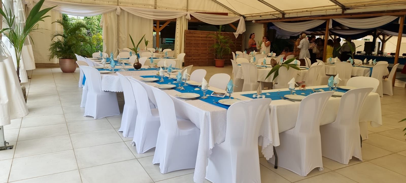 Un espace extérieur lumineux avec des tables nappées de blanc, des chemins de table bleus et des chaises recouvertes de housses blanches.