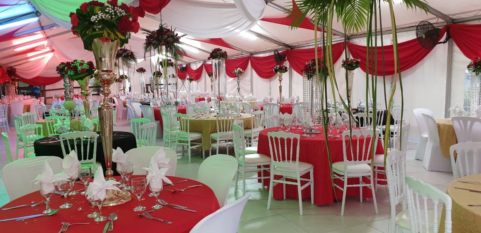 Une tente événementielle décorée de nappes rouges et or, de chaises blanches, de centres de table floraux et de draperies en tissu rouge.