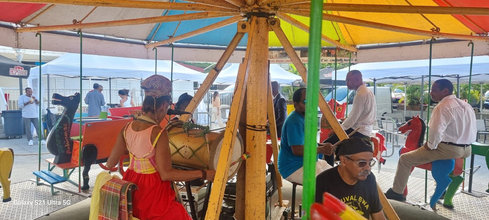 Des personnes sont assises sur un carrousel lors d'un événement en plein air, tandis qu'un musicien joue du tambour au centre, sous un dais coloré.