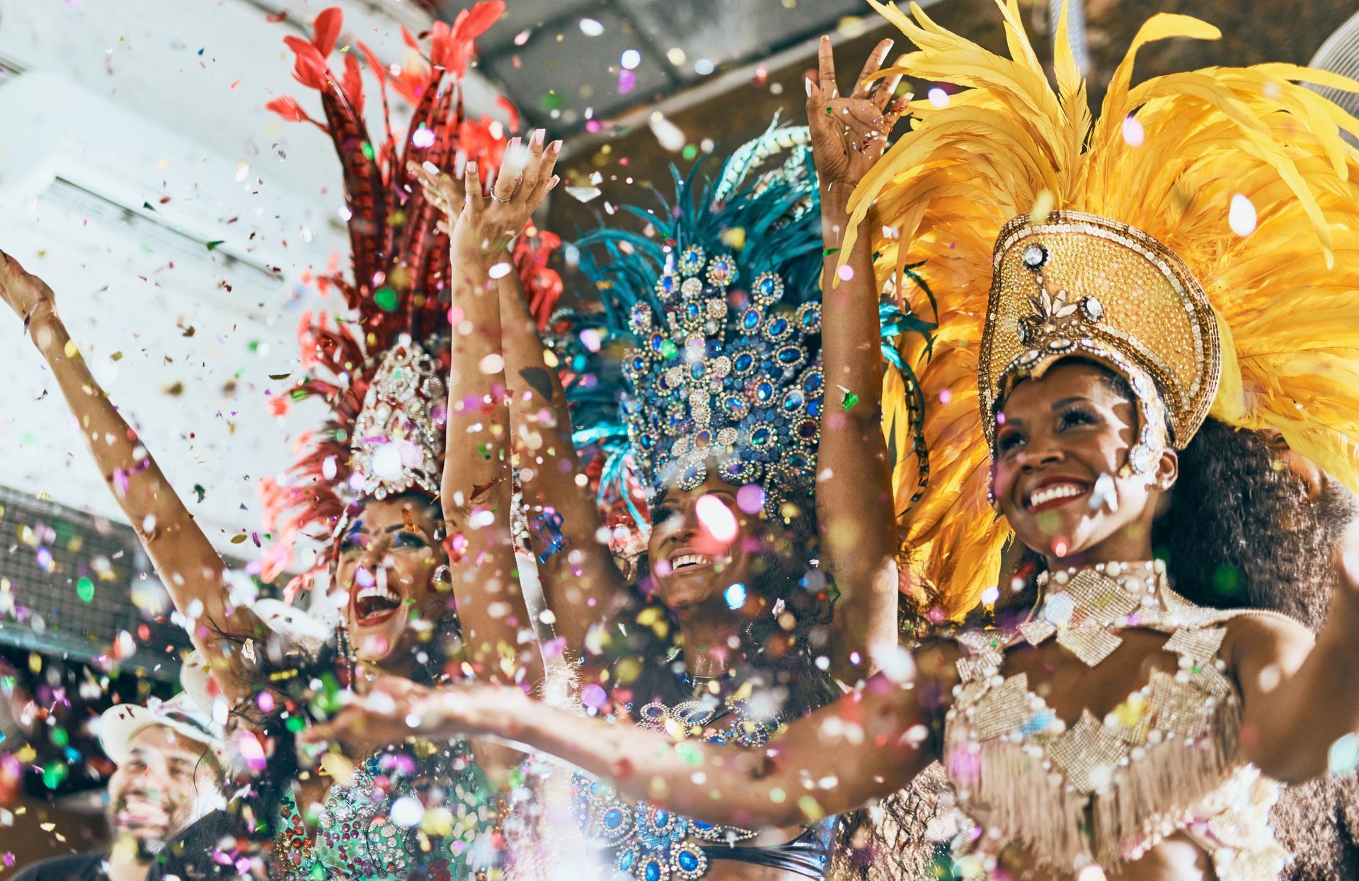 Trois artistes vêtus de costumes de carnaval élaborés et colorés et coiffés de parures de plumes posent les bras levés au milieu des confettis.