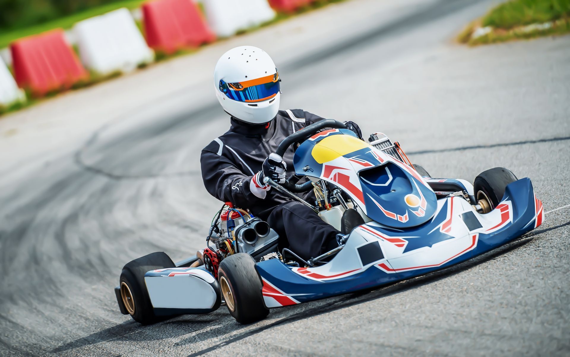 Une personne portant un casque et une combinaison de course conduit un kart bleu, blanc et rouge dans un virage sur une piste asphaltée.