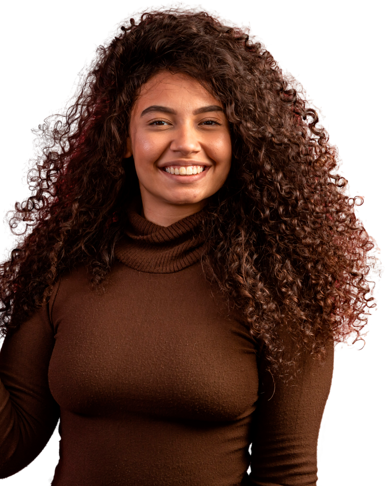 Une personne souriante aux cheveux foncés, volumineux et bouclés, portant un pull à col roulé marron sur fond blanc uni.