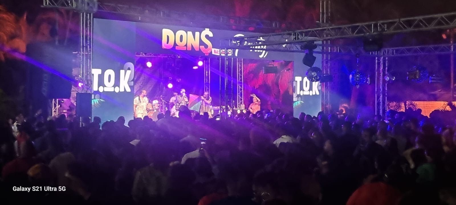 Une foule assiste à un spectacle en direct sur une scène éclairée de violet, où s'affichent les textes DONS et TOK.