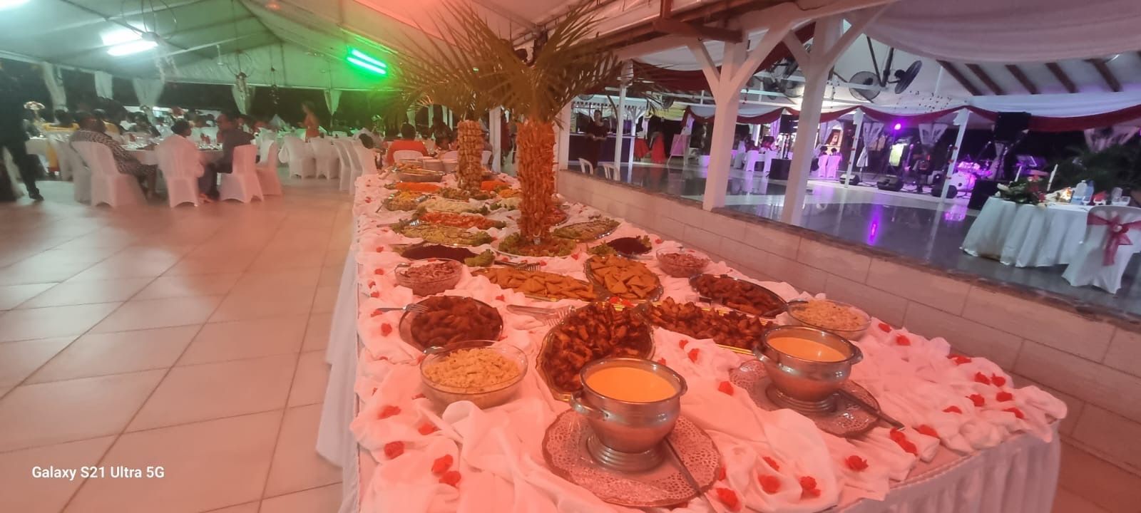 Une longue table de buffet garnie de plats variés dans une salle de banquet décorée et éclairée, avec des invités attablés.