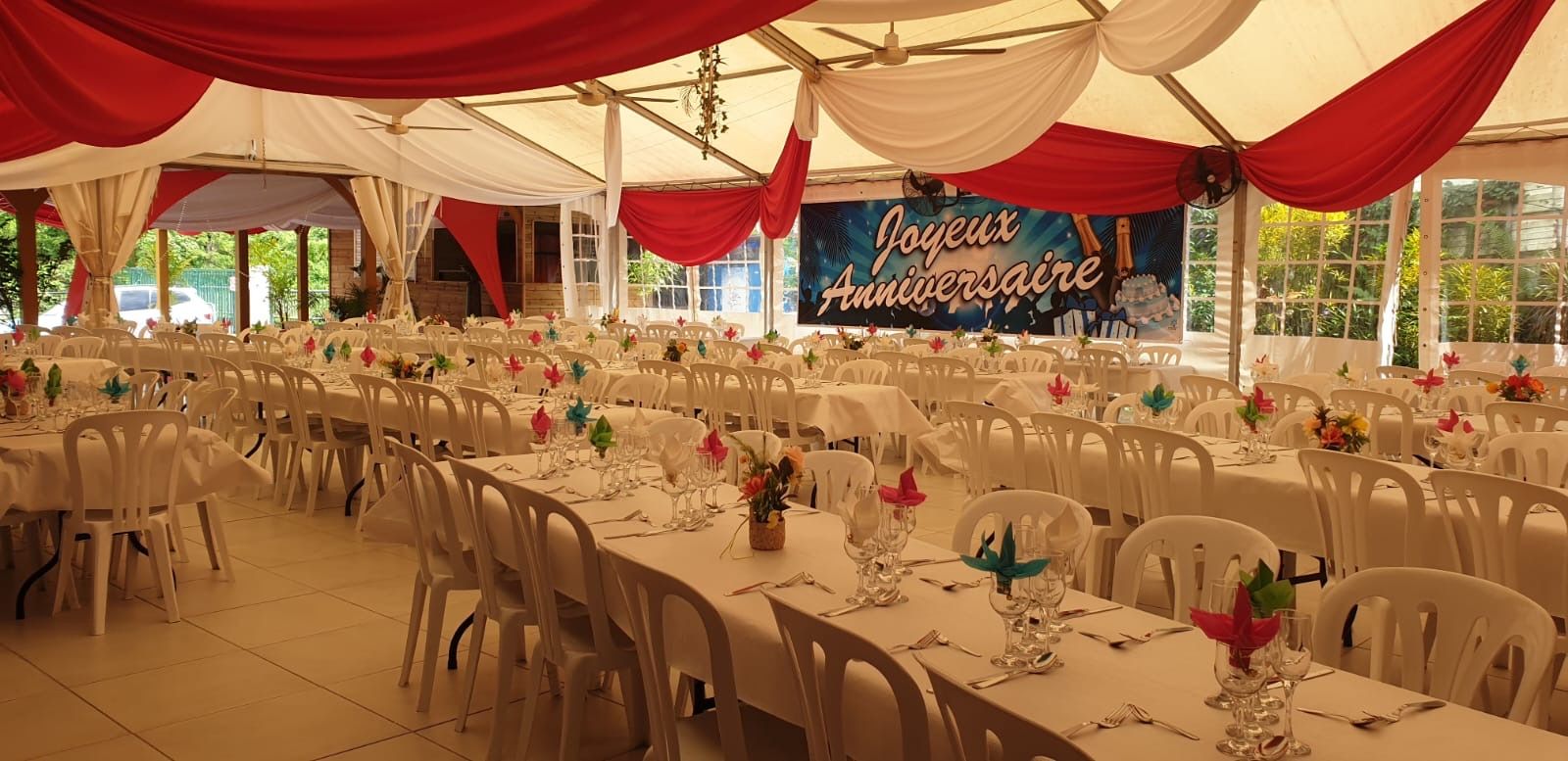 Une tente dressée pour une fête d'anniversaire, avec de longues rangées de tables, des chaises blanches, des tentures rouges et blanches et une banderole.