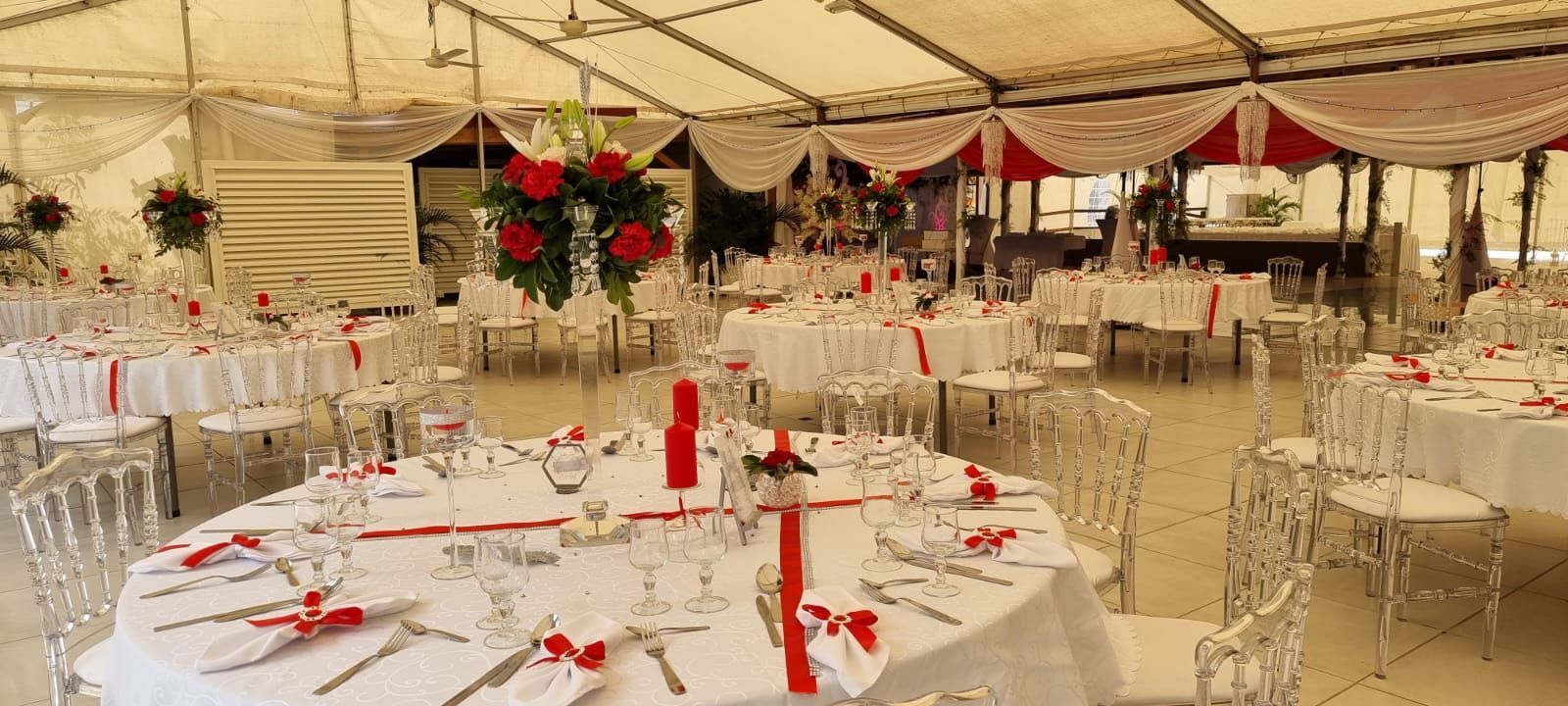 Une tente festive pour une réception de mariage, avec des tables rondes nappées de blanc, des touches de rouge et d'élégantes chaises transparentes.