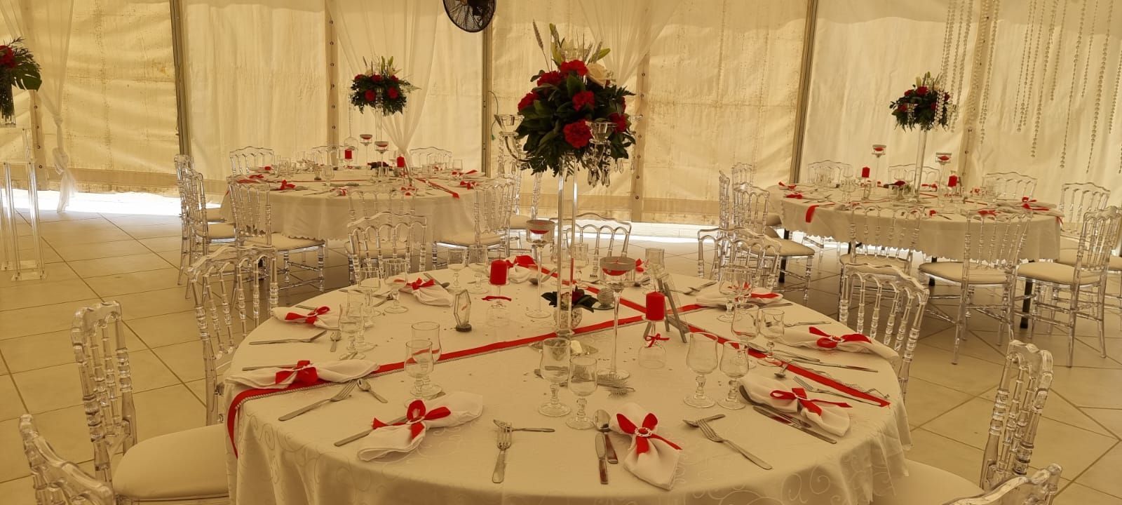 Une décoration pour une réception de mariage avec des tables rondes, du linge de table blanc, des chaises transparentes et des centres de table floraux agrémentés de touches de rouge.