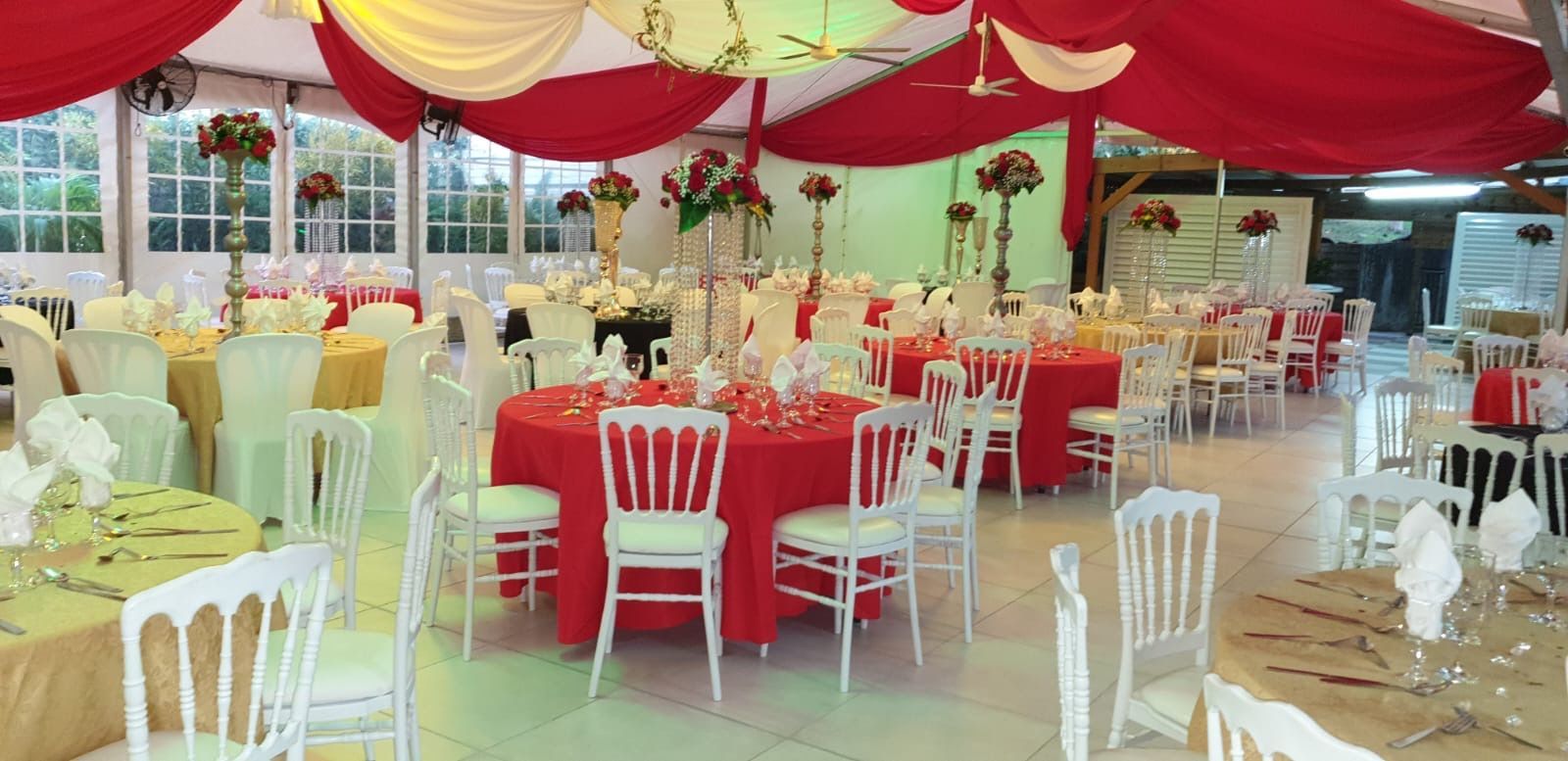Salle de réception décorée de tissus rouges et blancs, avec des tables rondes, des chaises blanches et des centres de table.