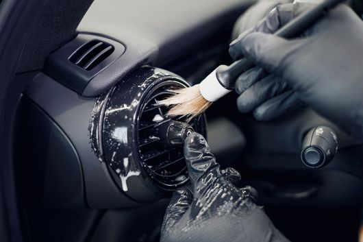 Des mains gantées nettoient la grille d'aération du tableau de bord d'une voiture avec un pinceau.