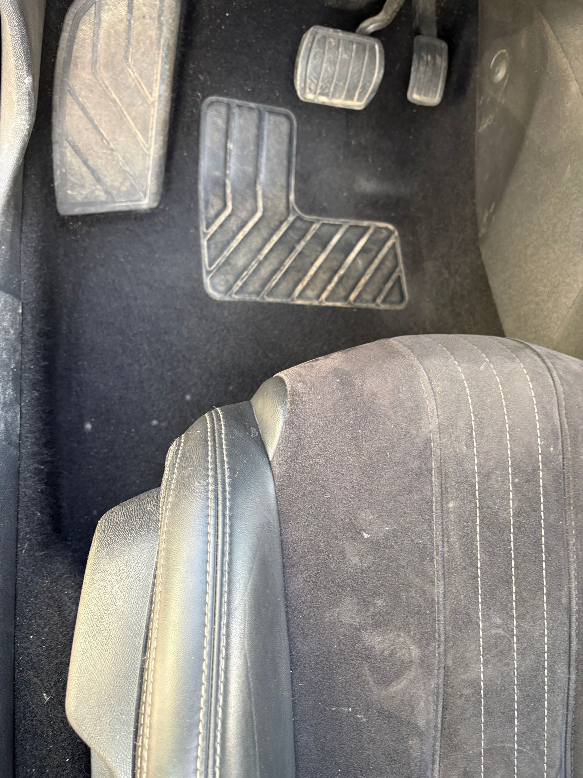 Plancher de voiture avec pédales noires et tapis de sol, visible à côté du siège avant.