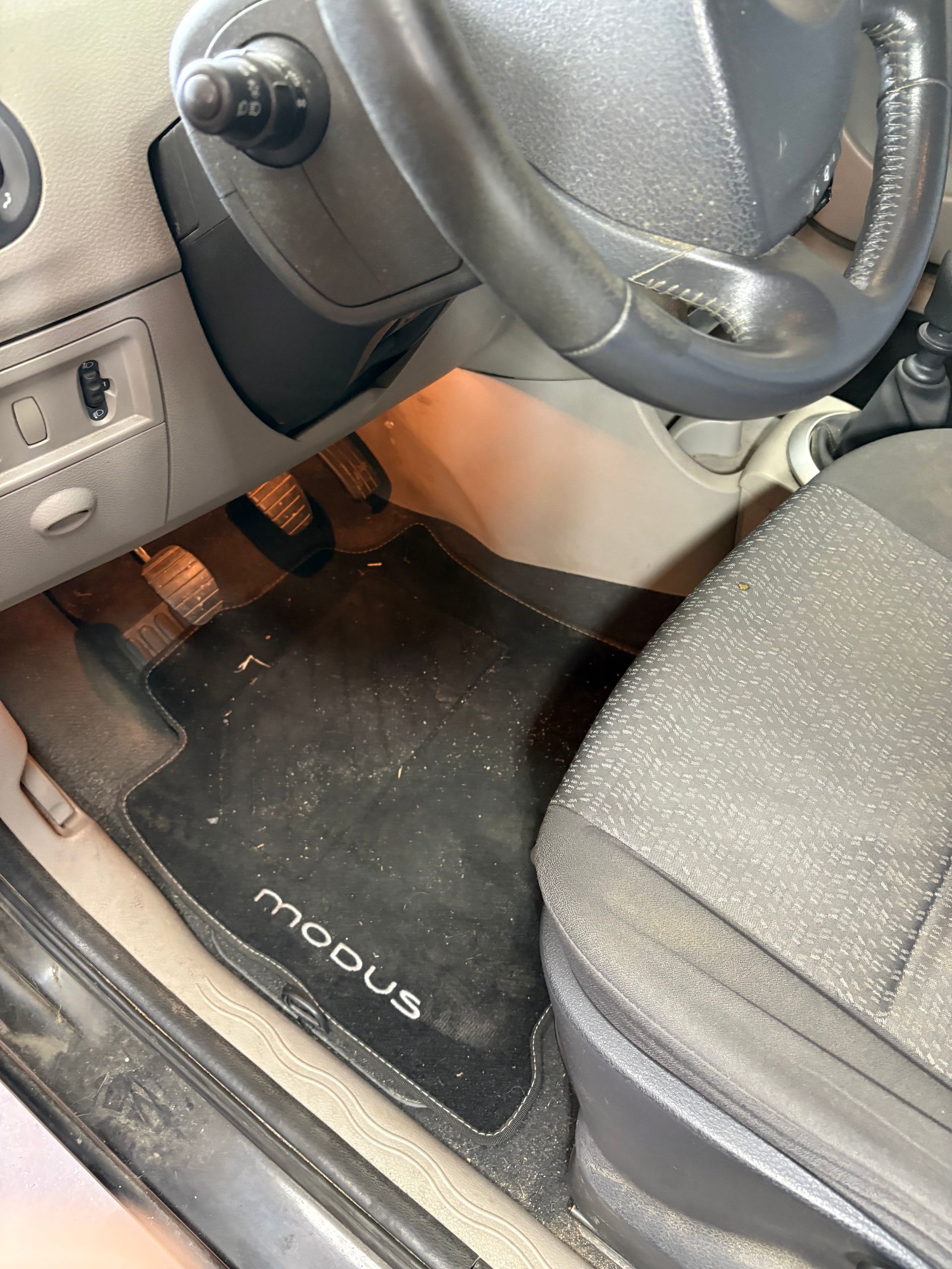 Intérieur d'une Renault Modus montrant le siège conducteur, le volant, les pédales et un tapis de sol noir.