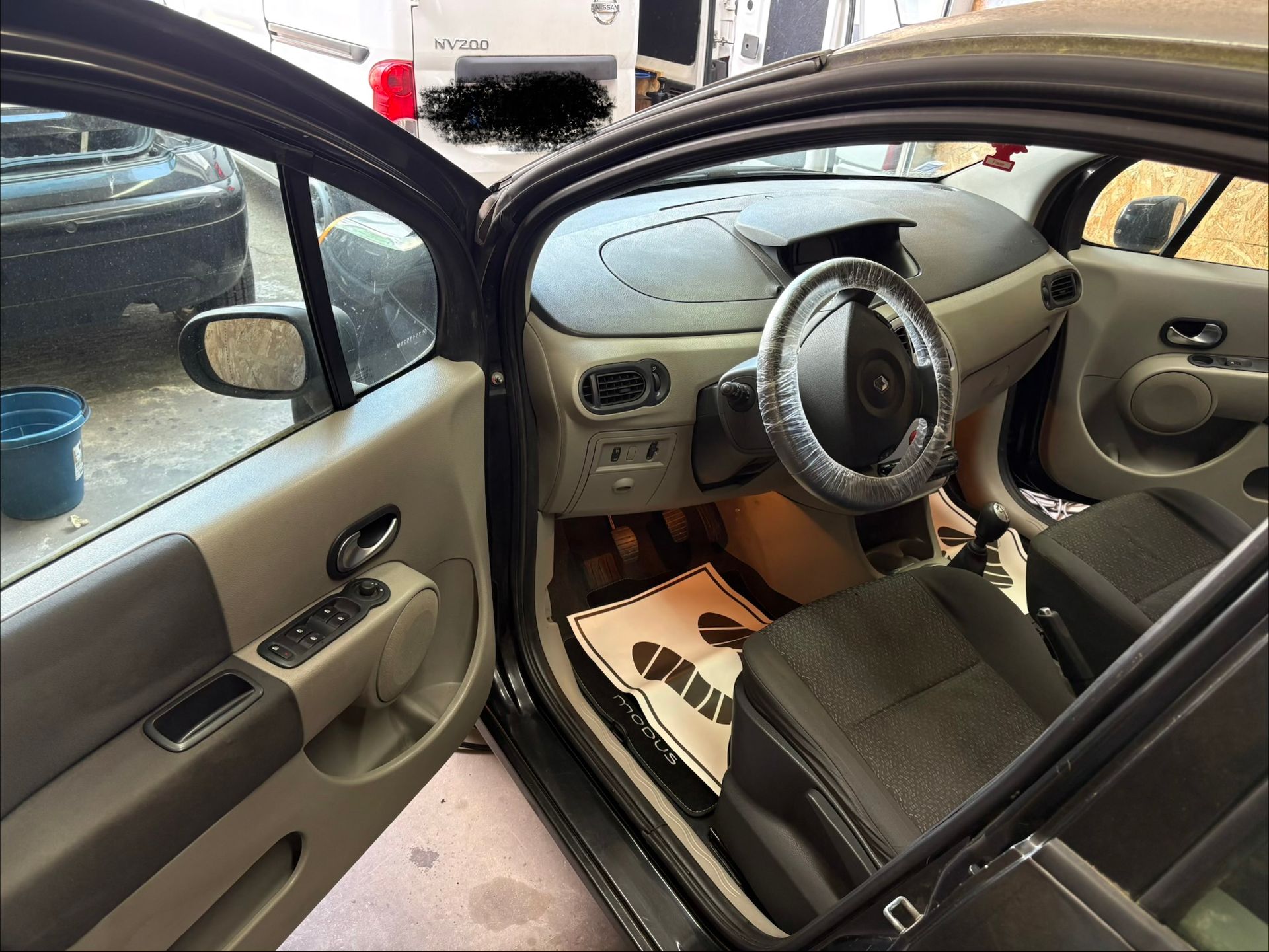 Portière de voiture ouverte, laissant apparaître un habitacle noir et beige, avec tableau de bord, volant et pédales.