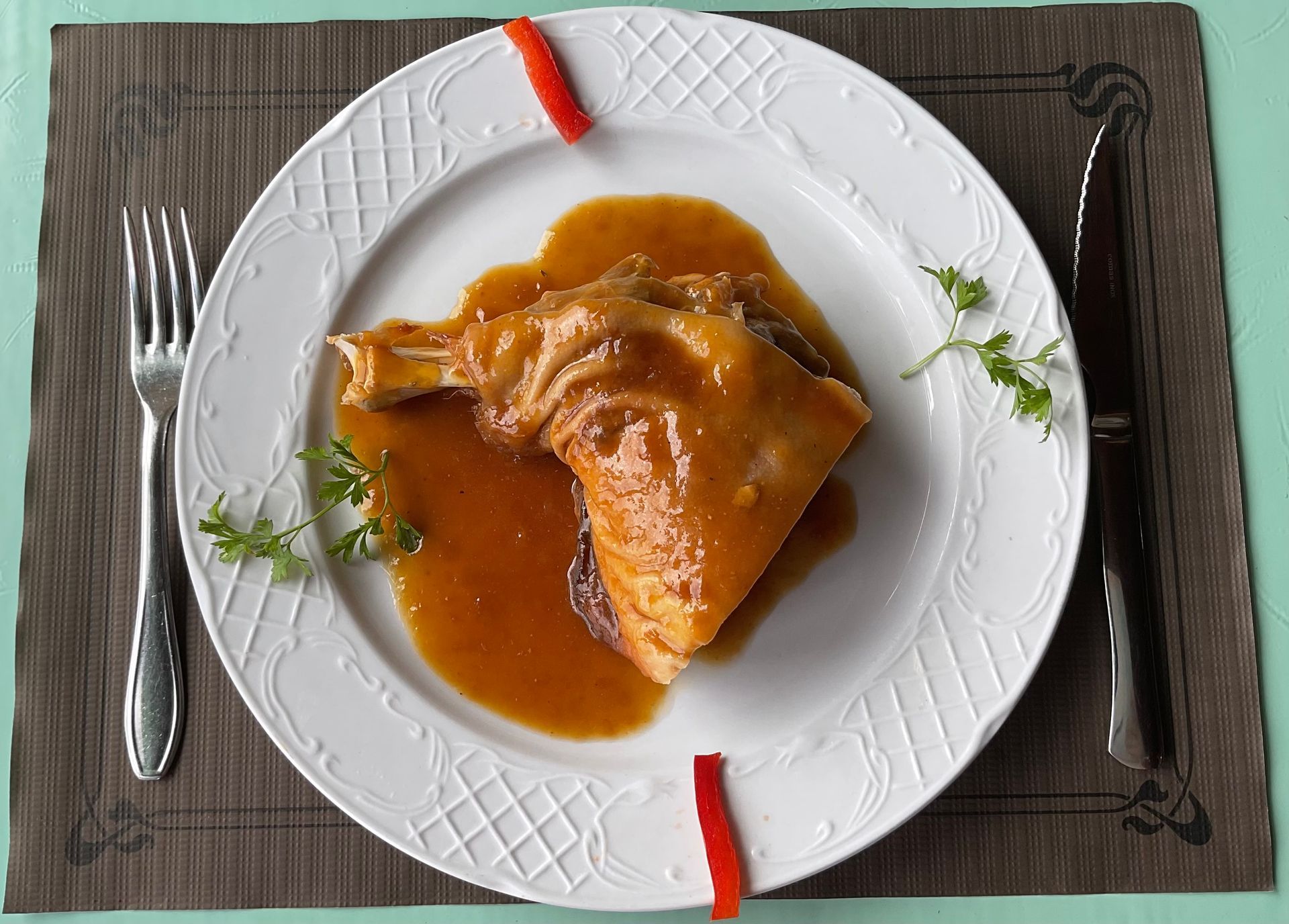 Pollo asado emplatado en salsa marrón, adornado con hierbas y pimiento rojo en un plato.