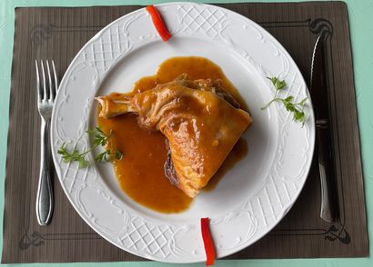 Pollo asado emplatado en salsa marrón, adornado con hierbas y pimiento rojo en un plato.