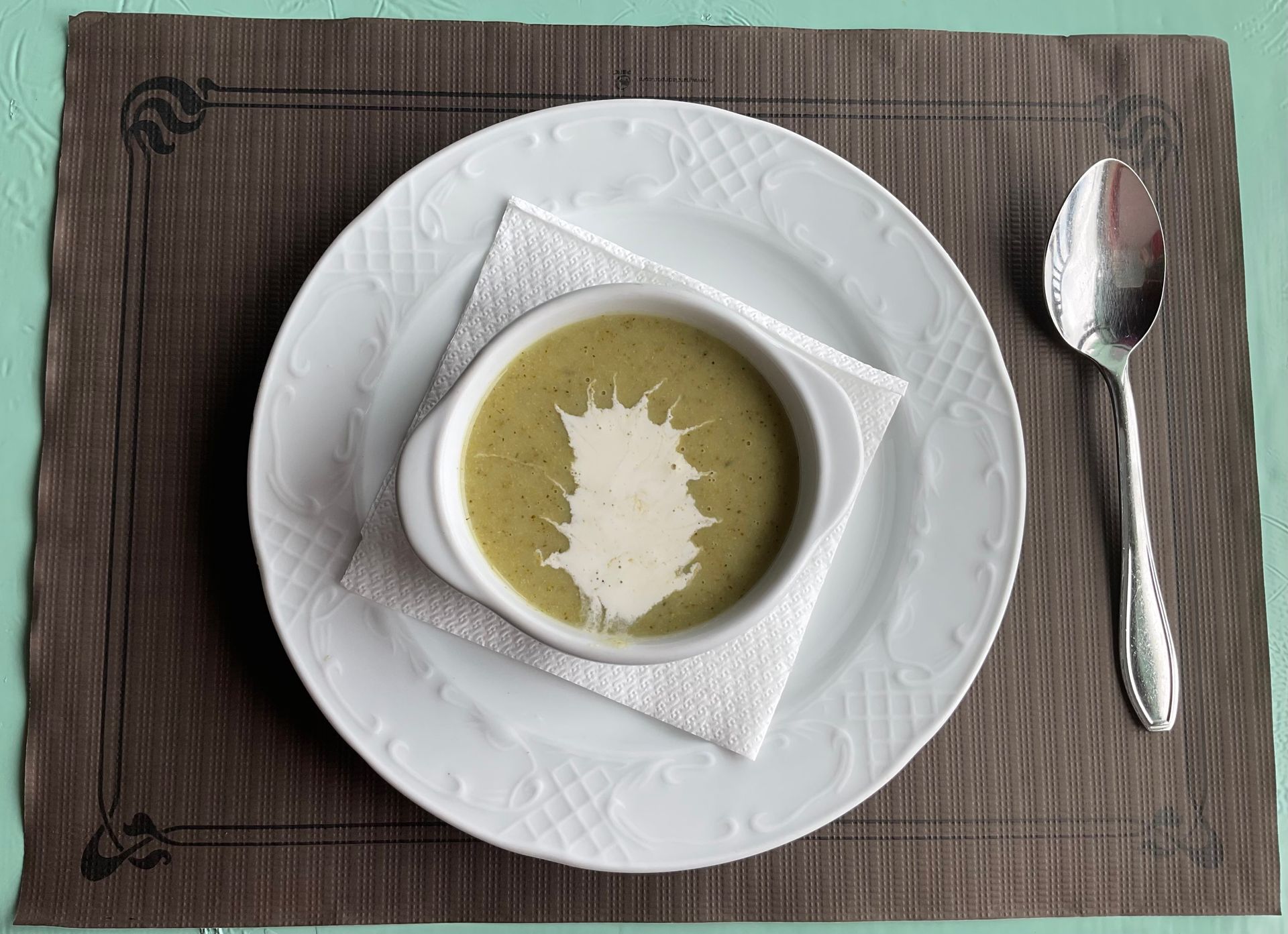 Sopa servida en un tazón blanco sobre un plato con una cuchara, todo colocado sobre un mantel individual marrón.