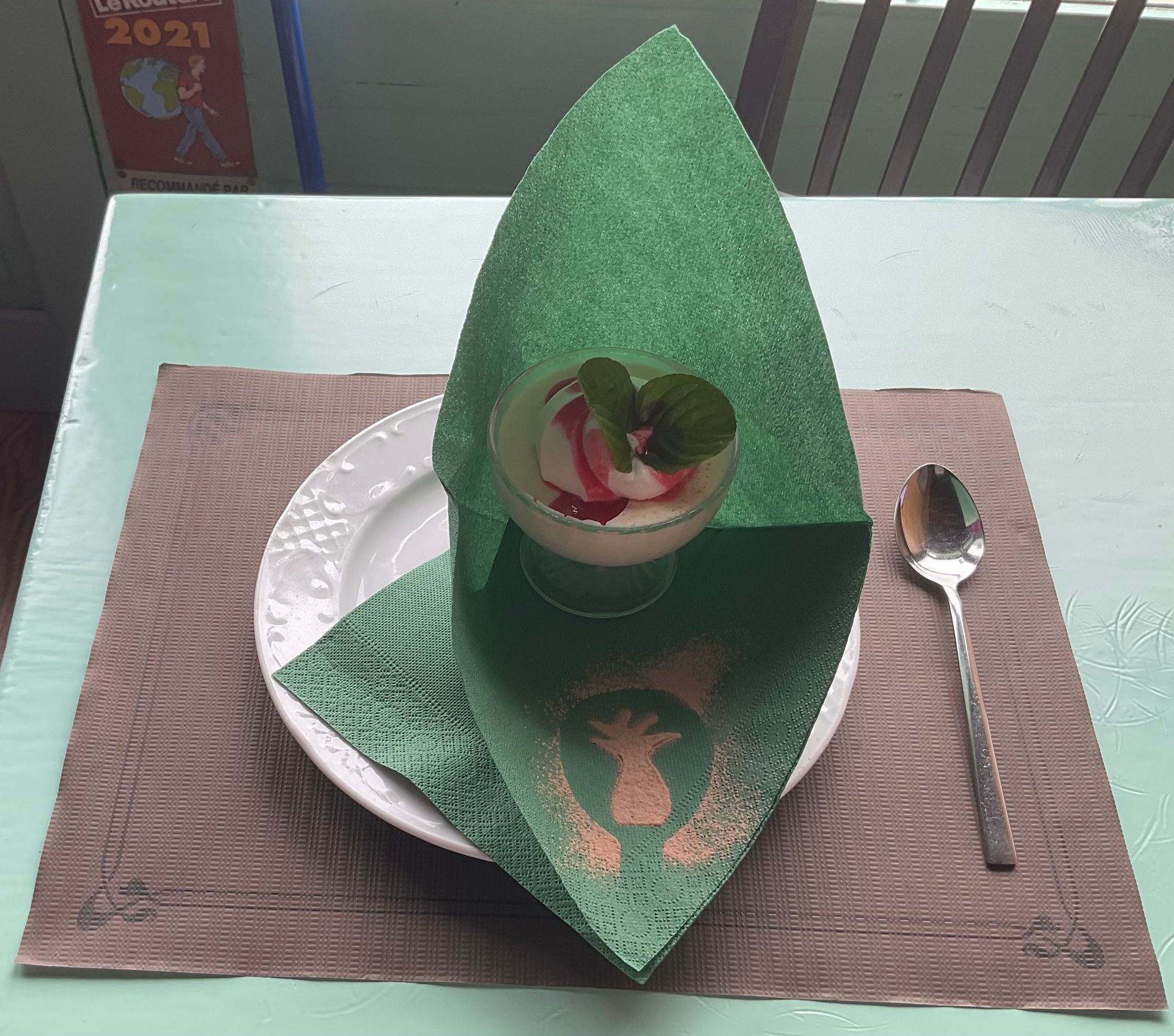 Servilleta verde doblada sobre un plato blanco con postre, sobre un mantel individual marrón al lado de una cuchara.