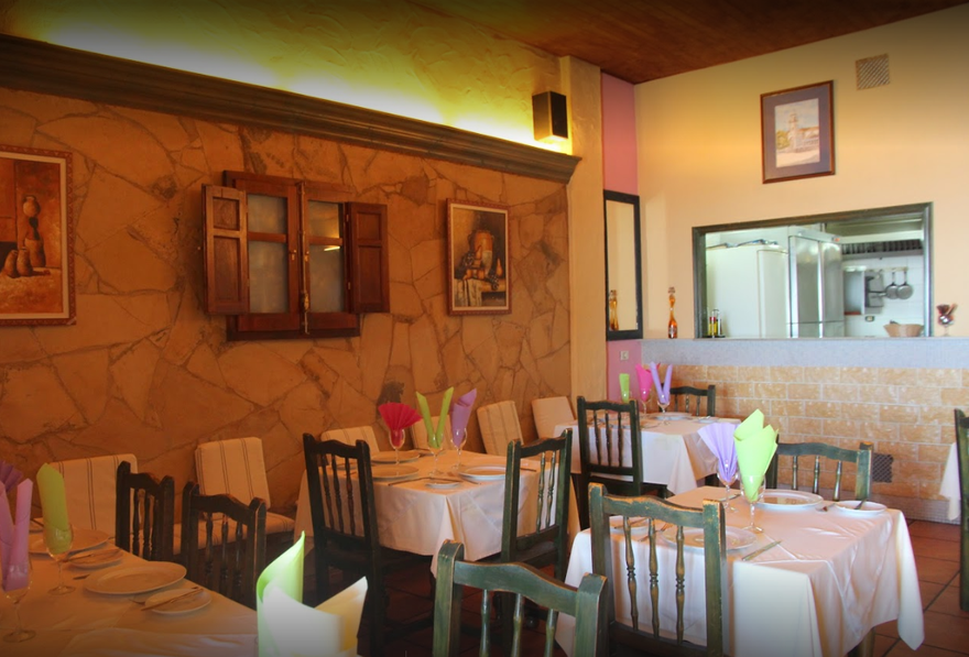 Comedor de restaurante con iluminación cálida, mesas vestidas con manteles blancos y sillas junto a paredes de piedra y un espejo.