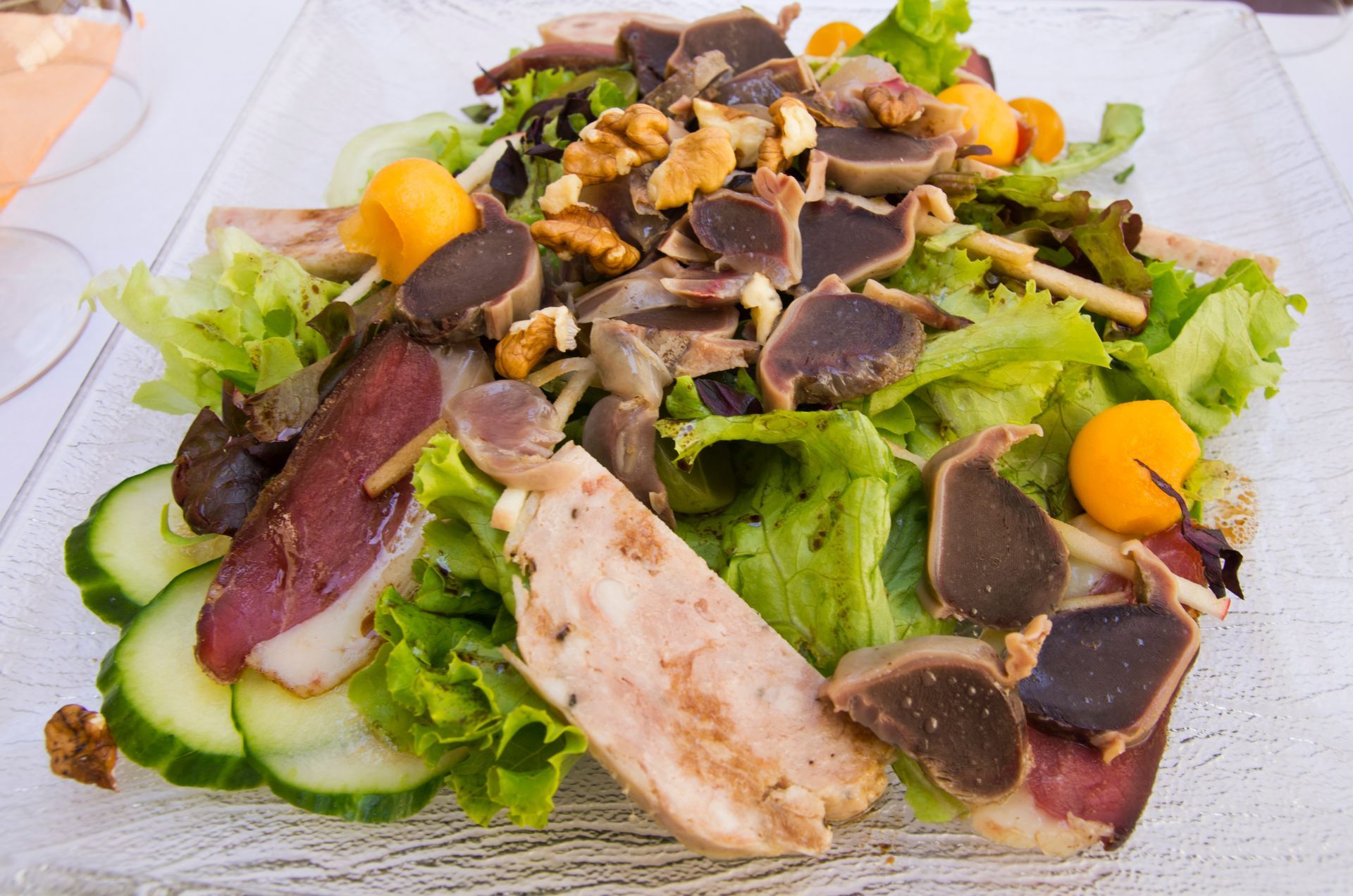 Une assiette transparente comprenant des gésiers, du magret de canard, salade, noix, jambon de pays