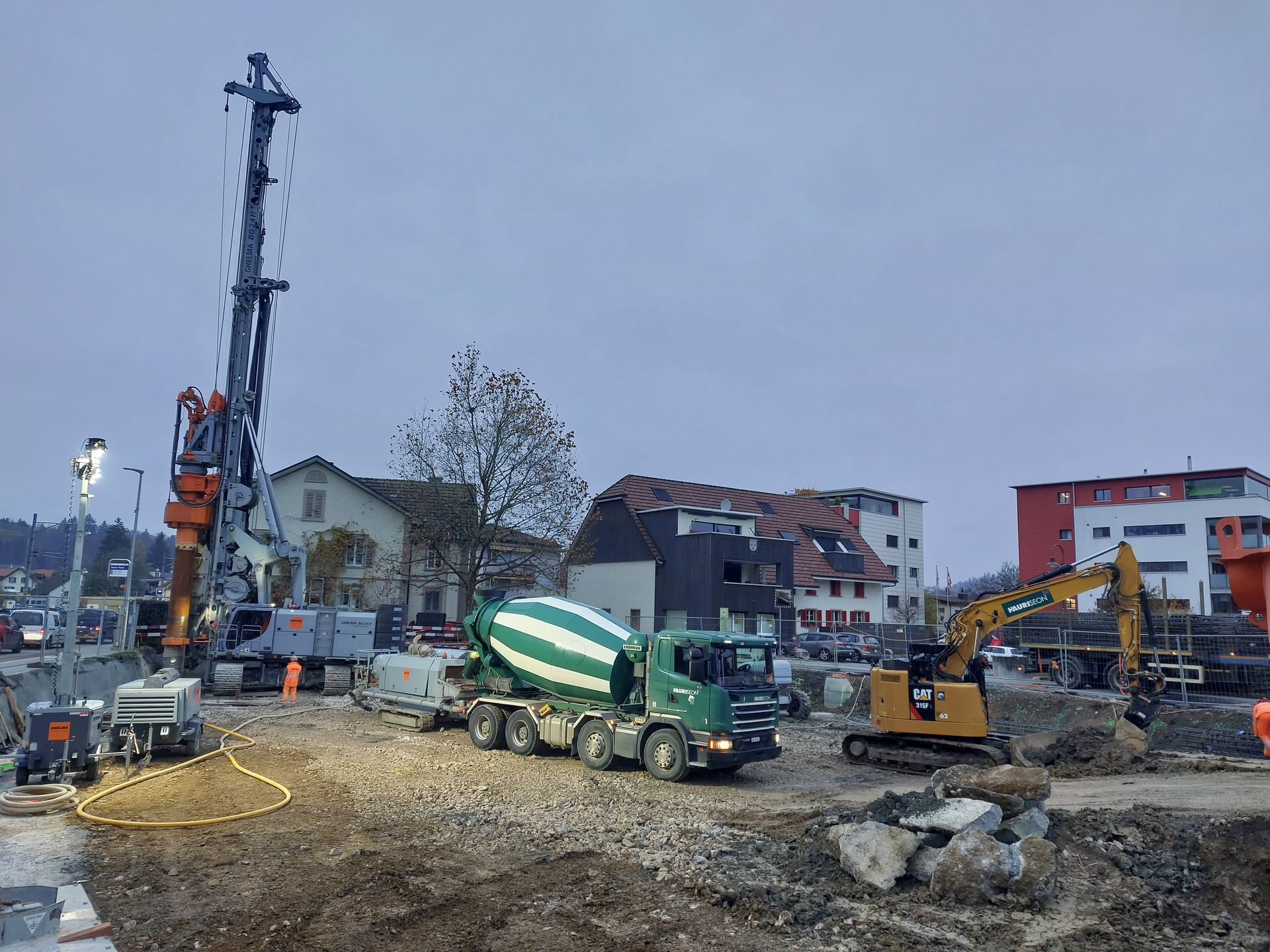 Baustelle mit Bohrmaschine, Zementlaster und Bagger; bewölkter Himmel.