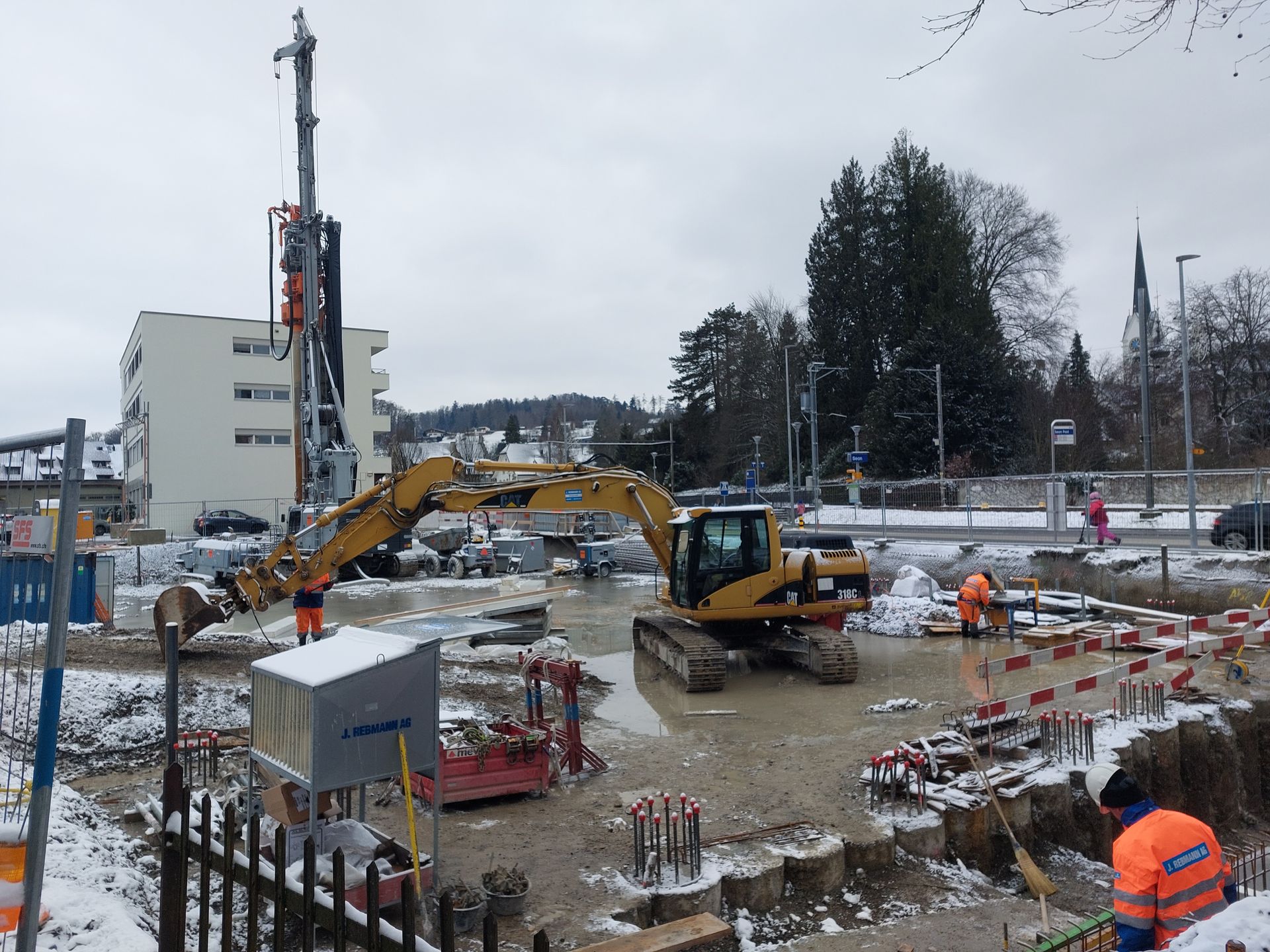 Baustelle mit Bagger und Bohrgerät; Arbeiter in orangefarbenen Westen; schneebedeckter Boden; Gebäude und eine Kirche im Hintergrund.
