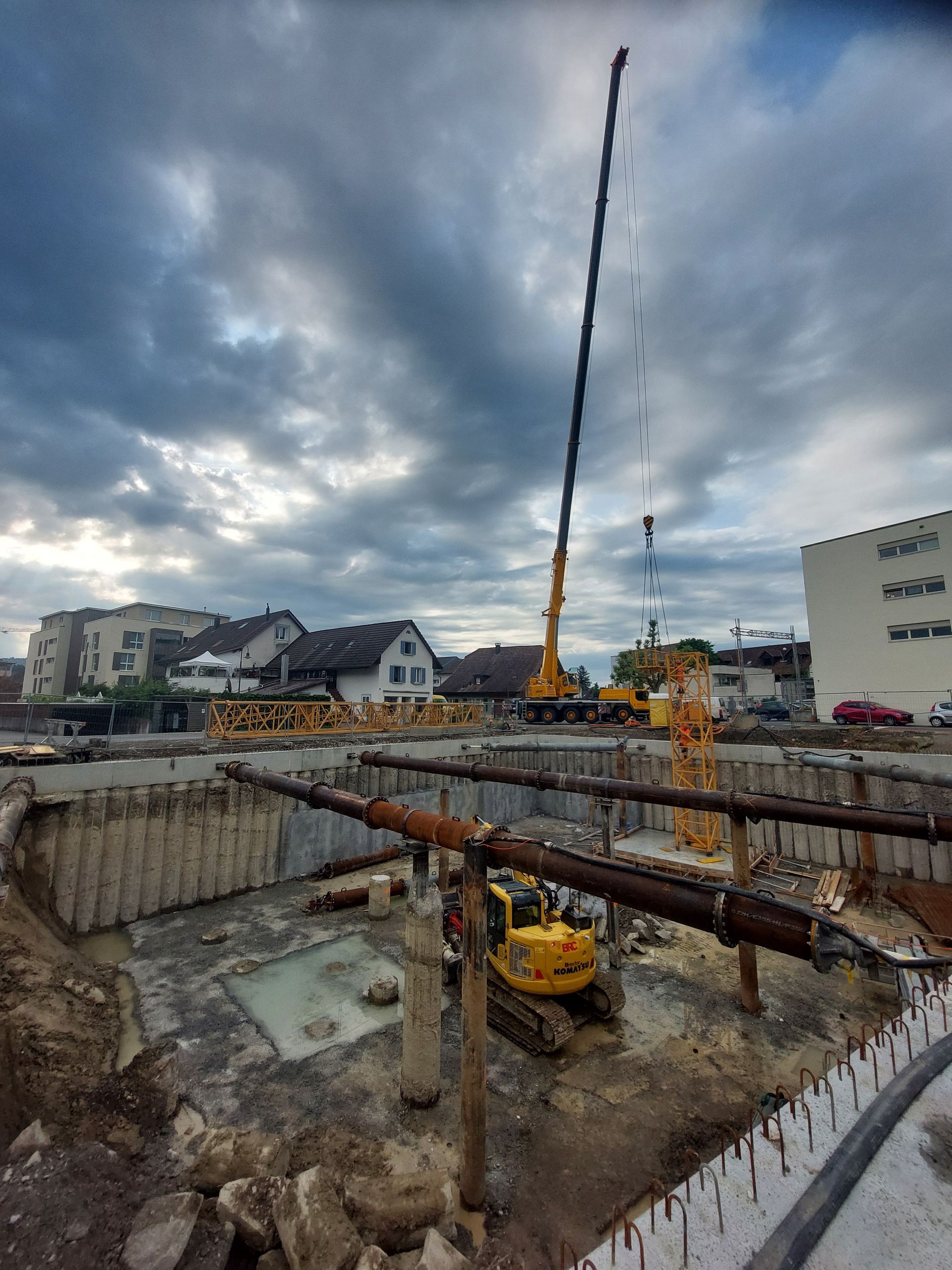 Baustelle mit hohem Kran, Bagger und tiefer Baugrube unter bewölktem Himmel.