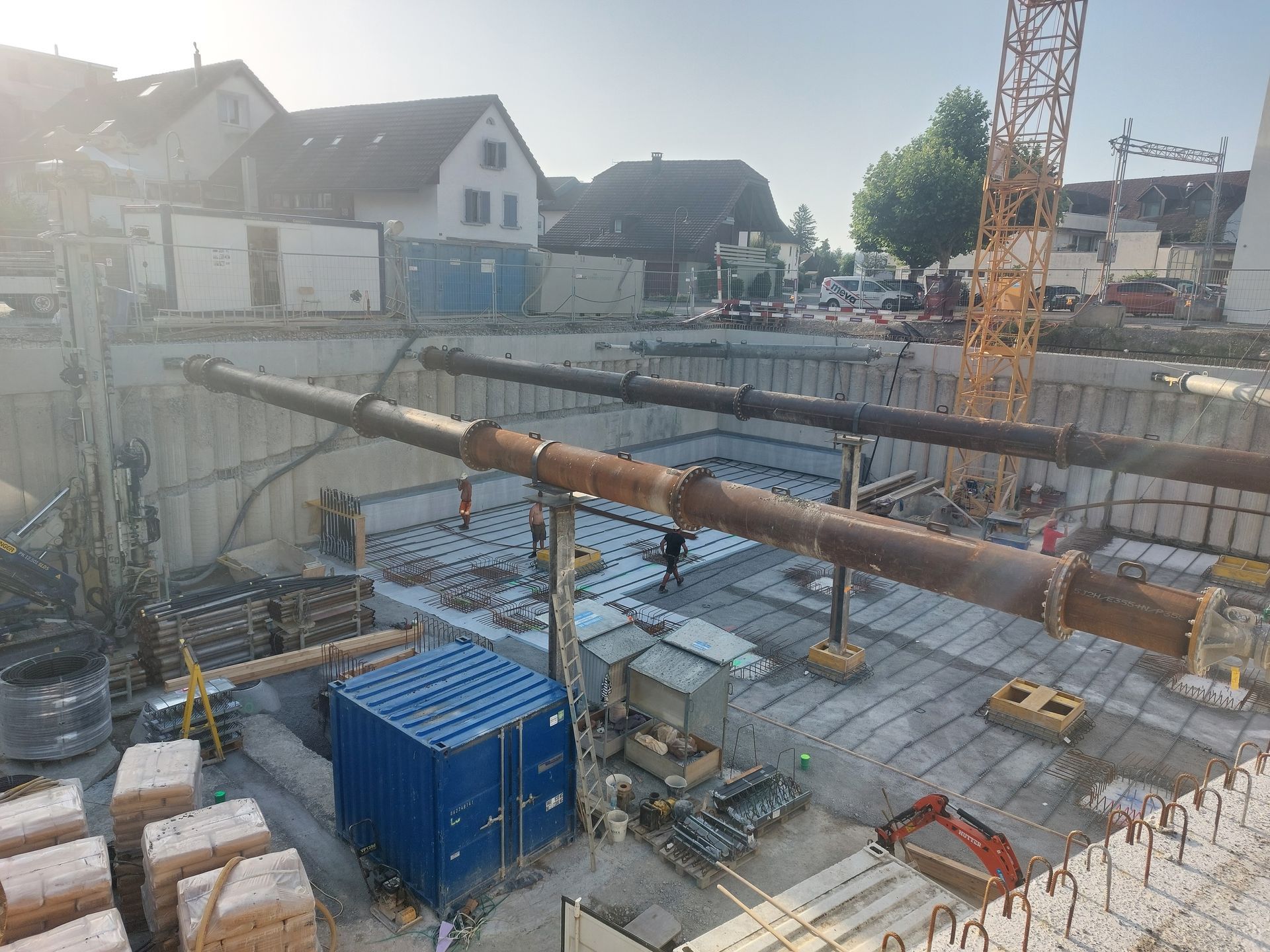 Baustelle mit großen Rohren über Betonfundament, Kran und Gebäuden im Hintergrund.