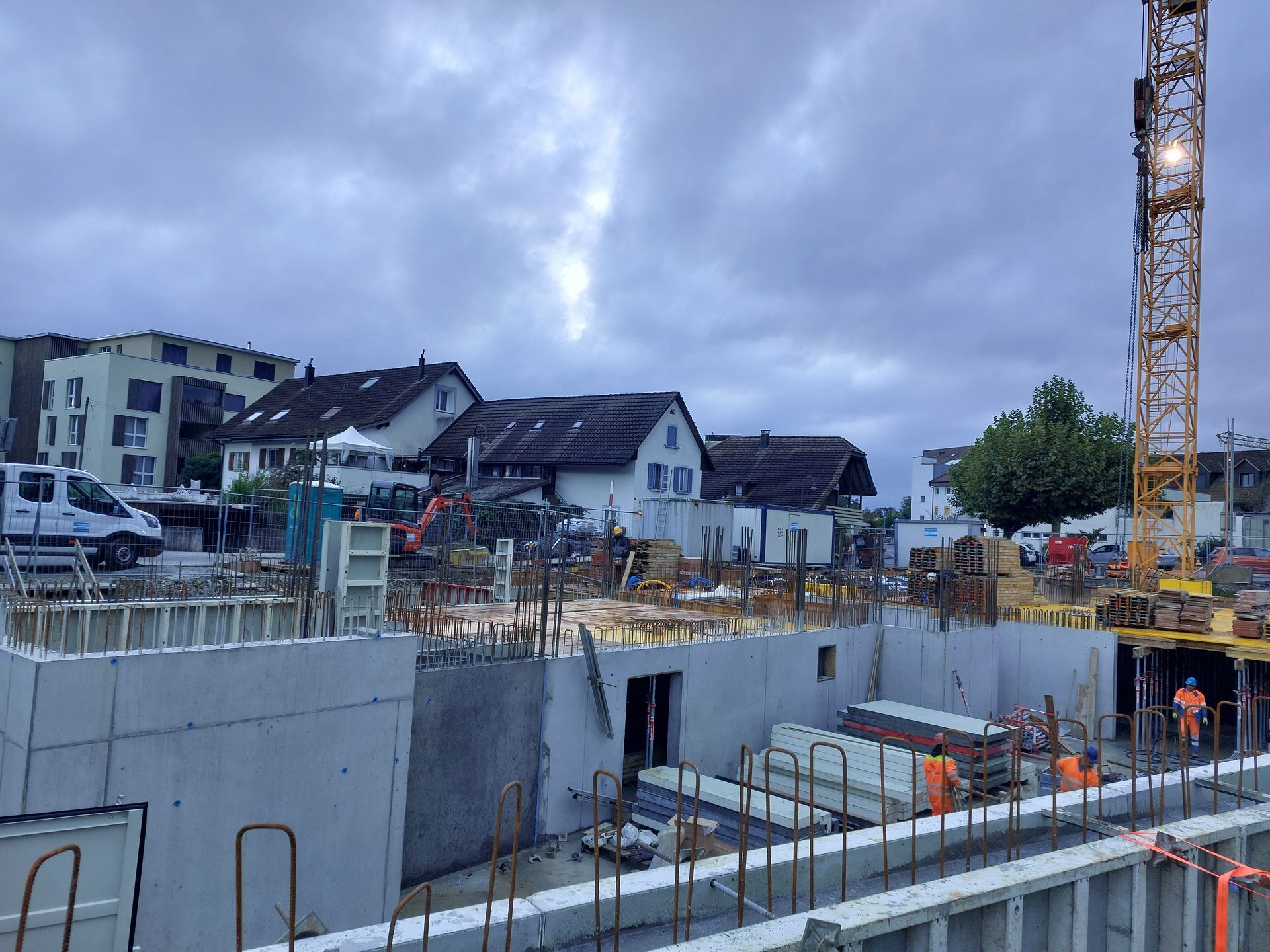Baustelle mit Kran, Betonwänden und Arbeitern. Im Hintergrund Häuser unter bewölktem Himmel.