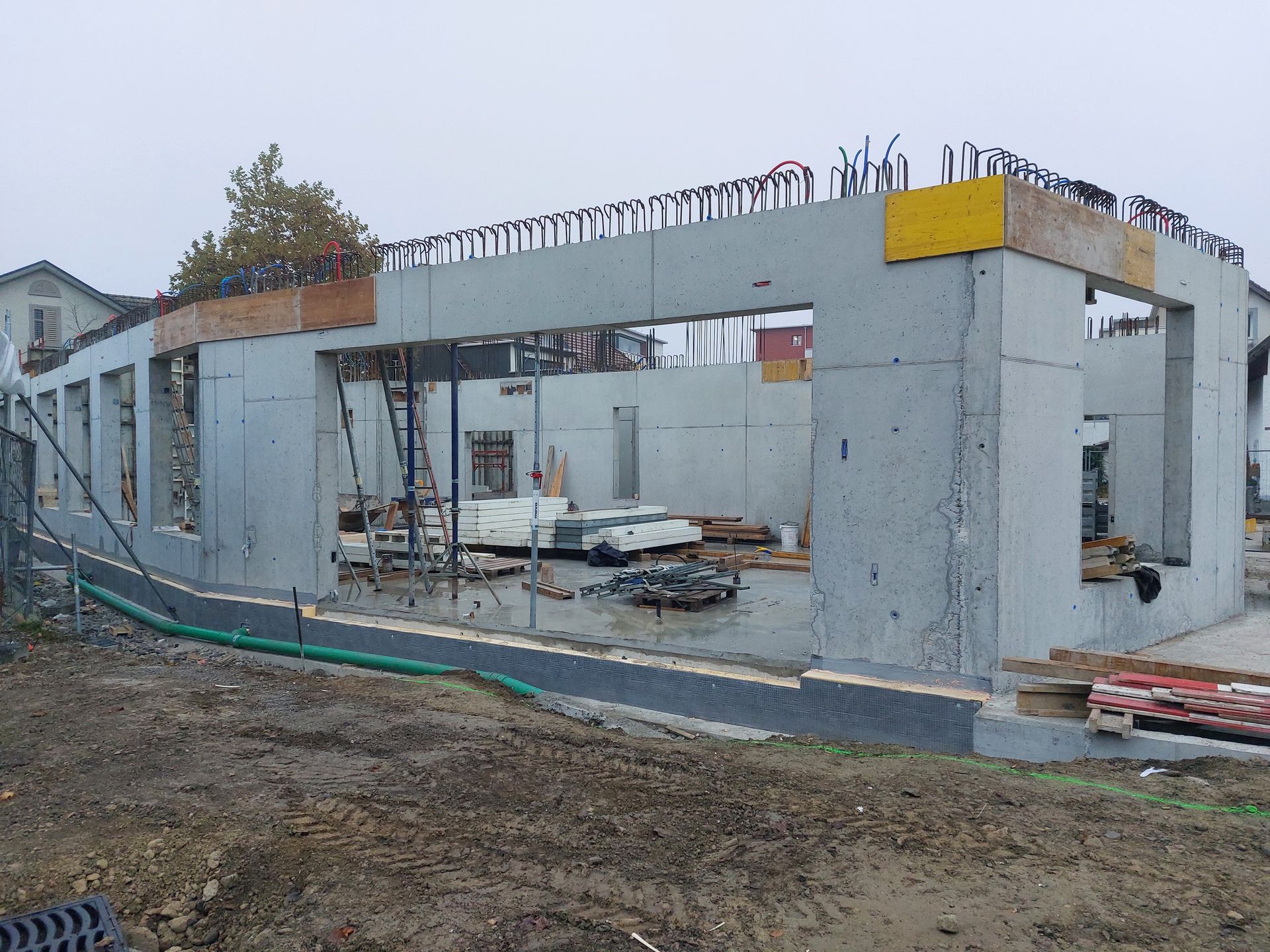 Baustelle: Betongebäude mit freiliegender Bewehrung; Grau- und Erdtöne; bedeckter Himmel.