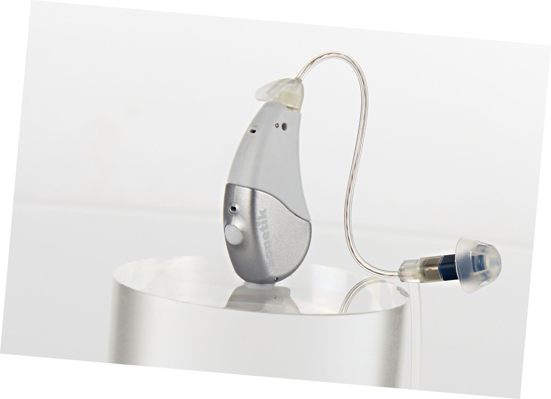 Silbernes und graues Hörgerät auf einem durchsichtigen Ständer; durchsichtiger Schlauch und Ohrstück.