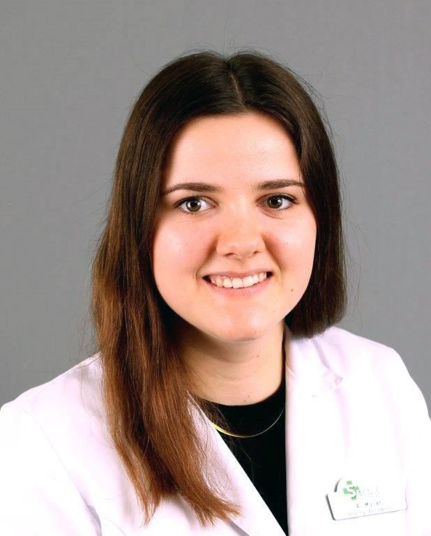 Frau mit langen braunen Haaren, trägt einen weißen Laborkittel und lächelt in die Kamera.