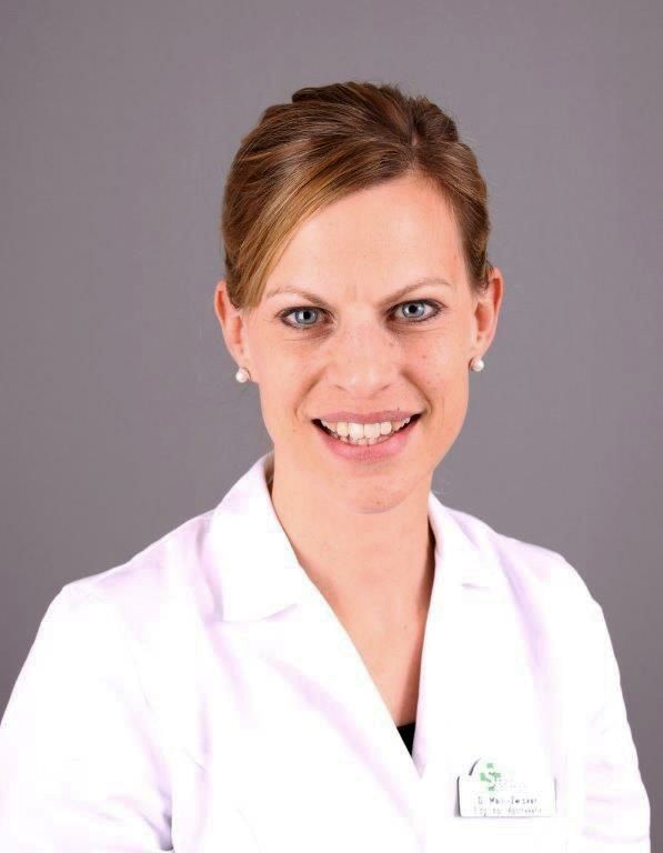 Frau im weißen Laborkittel, lächelt in die Kamera. Braunes Haar hochgesteckt, Perlenohrringe, grauer Hintergrund.