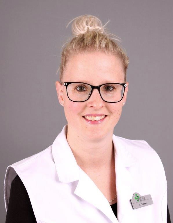 Frau mit blondem Dutt, Brille und weißer Uniform lächelt, wahrscheinlich in einer Apotheke.