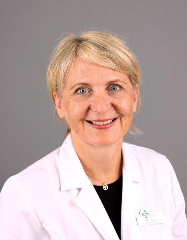 Frau im weißen Laborkittel, Brille, lächelnd. Grauer Hintergrund.