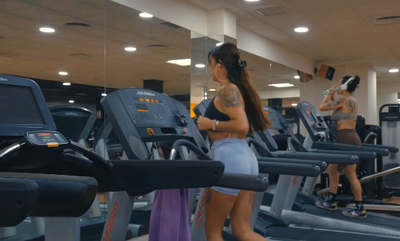 Mujer corriendo en una cinta de correr en un gimnasio, otra persona corriendo en el fondo.