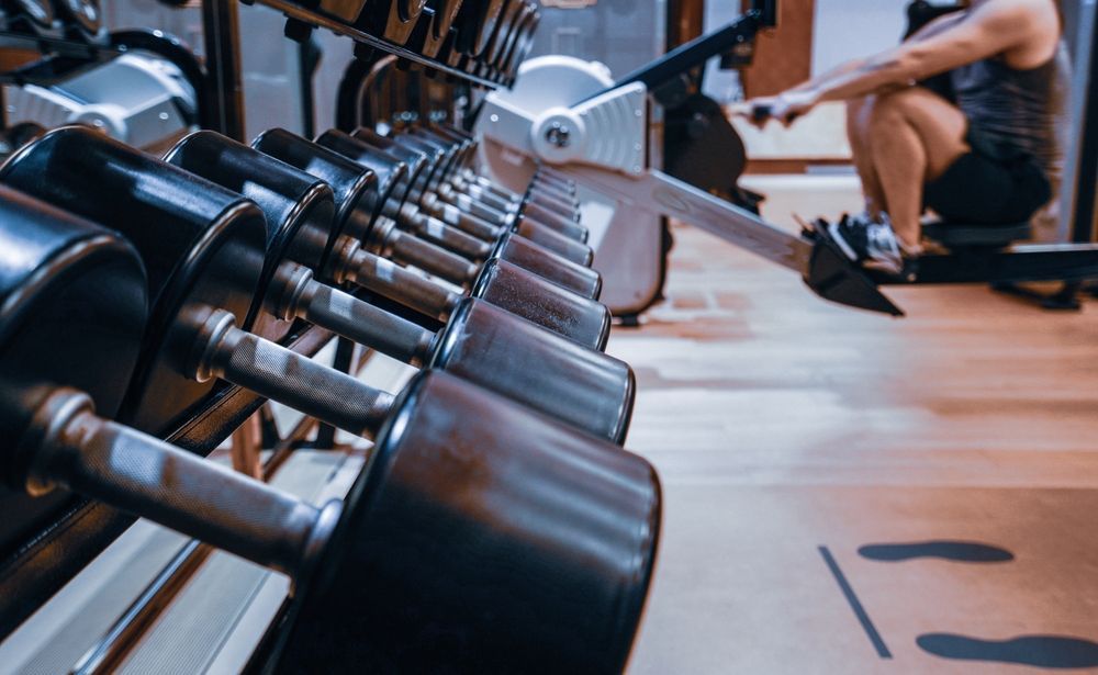 Fila de mancuernas en un gimnasio, con una persona usando una máquina de remo en el fondo.