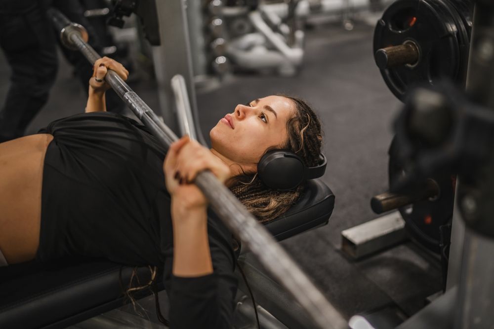 Mujer levantando pesas en un banco de press de banca en un gimnasio, usando auriculares.