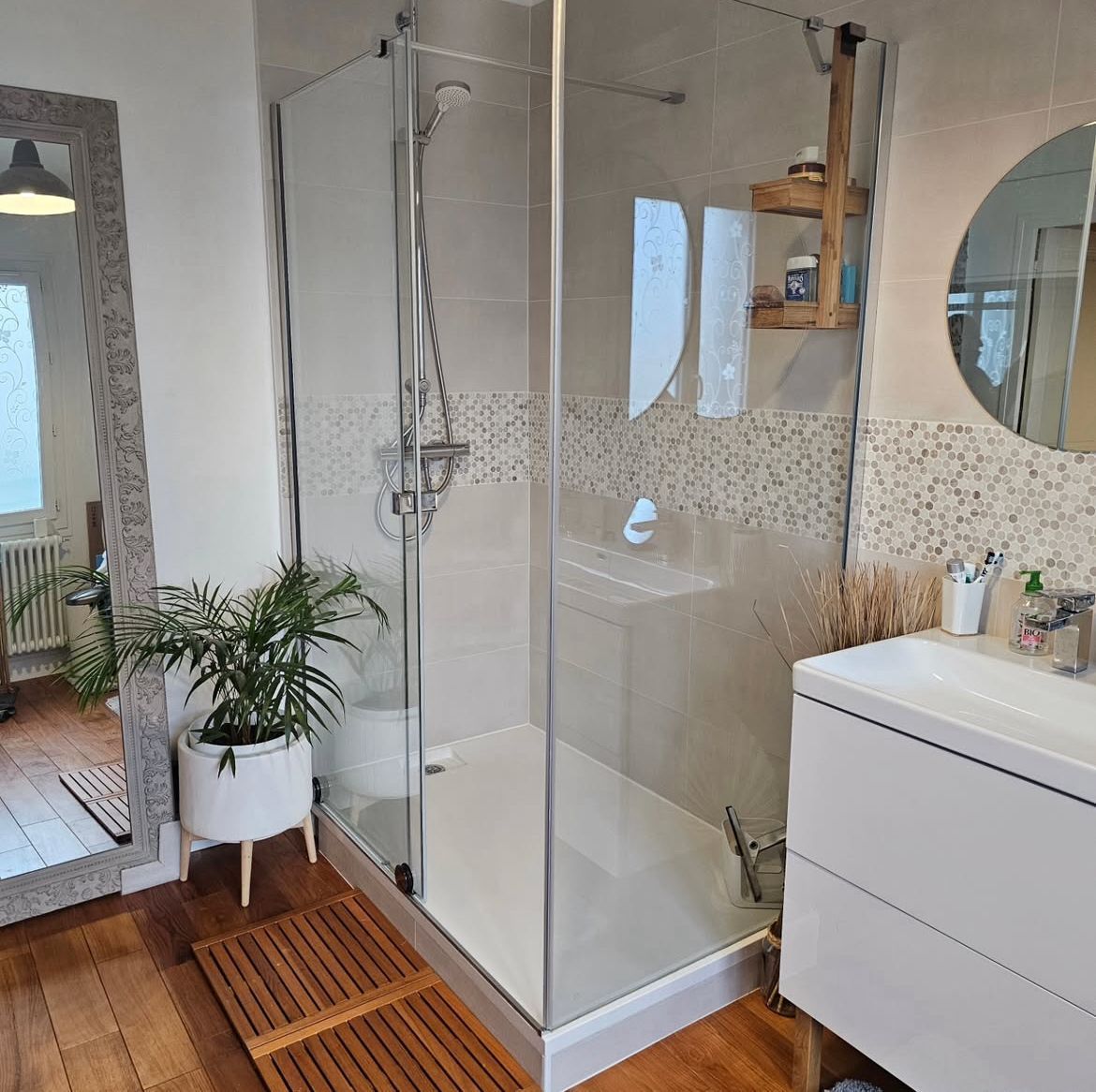 Une salle de bains moderne avec une douche vitrée, un parquet, un meuble-lavabo blanc, un miroir rond et une plante en pot.