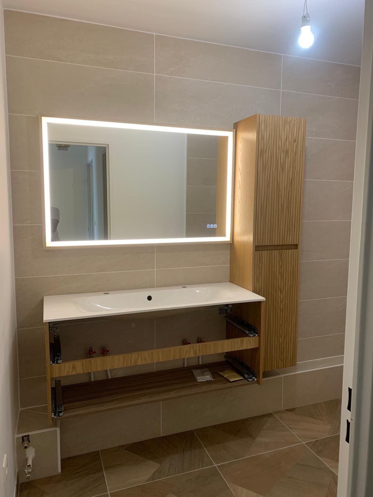 Un meuble-lavabo avec miroir lumineux mural et armoire latérale en bois, le tout sur un mur carrelé beige.