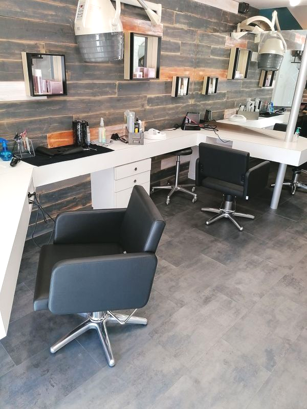 Intérieur d'un salon de coiffure avec postes de coiffage, fauteuils et miroirs. Mur en bois foncé, comptoirs blancs et sol gris.