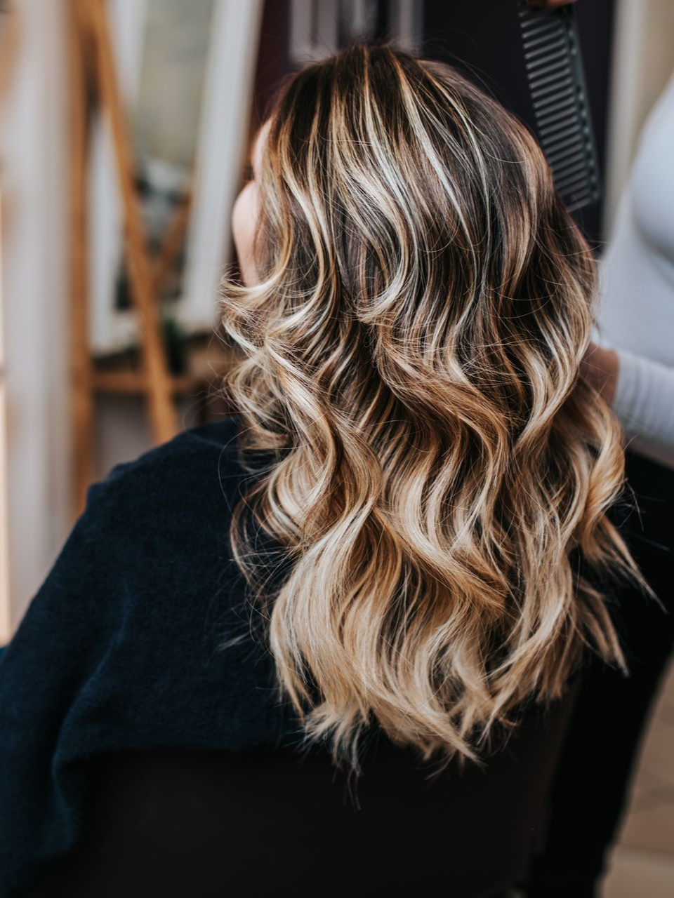Une femme aux cheveux ondulés et méchés est assise sur une chaise pendant qu'un coiffeur les peigne.
