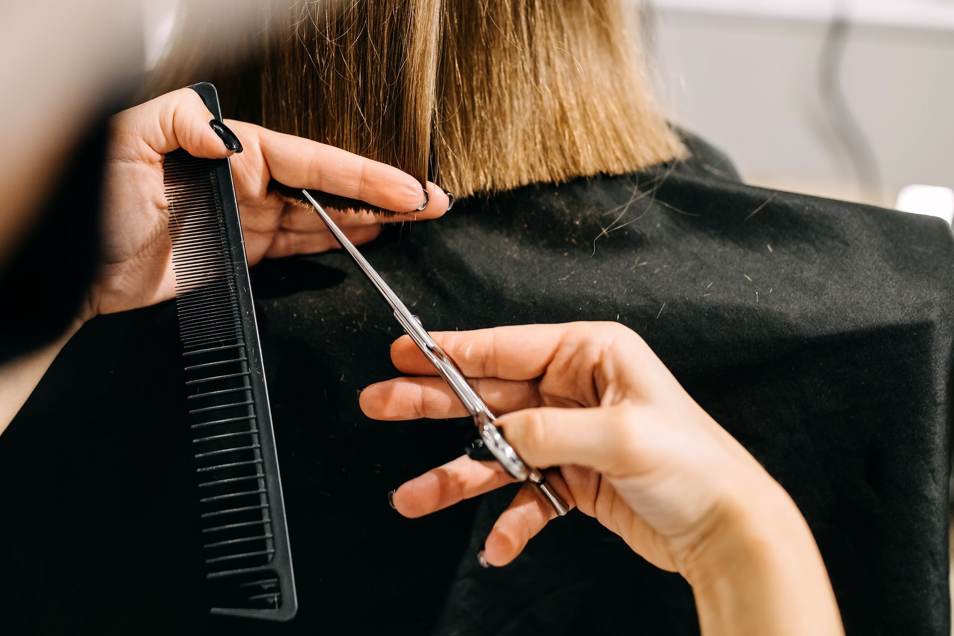 Des mains tenant un peigne et des ciseaux, coupant des cheveux blonds, une personne portant une cape noire dans un salon de coiffure.